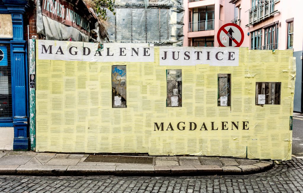 A large yellow billboard sits on a concrete sidewalk next to an intersection. Across the top of the billboard “Magdalene Justice” is printed in black text.