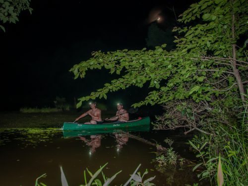 Two shirtless men wearing headlamps row a green canoe through a pond in otherwise pitch-black darkness.