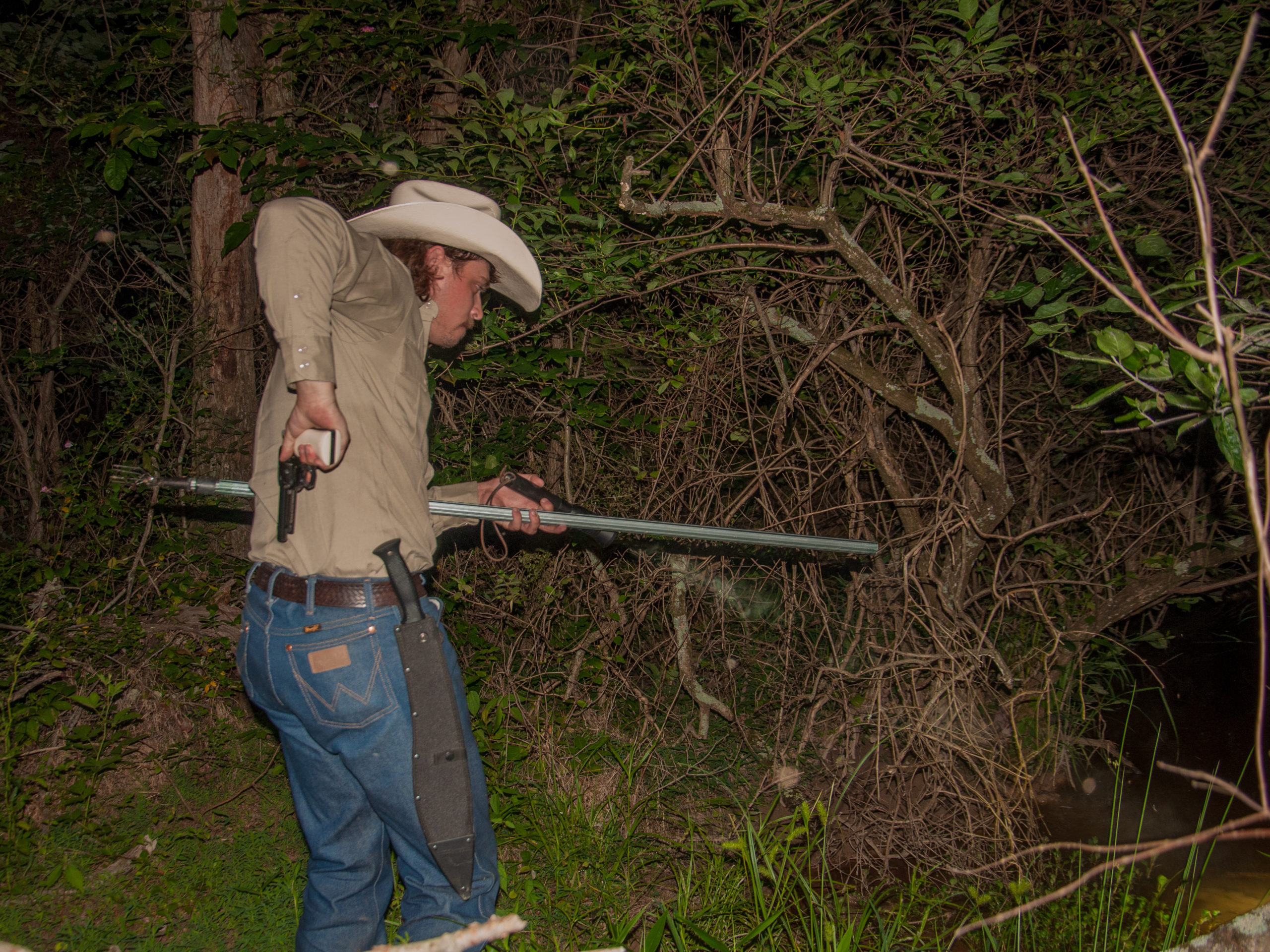 In the woods, a man wearing a tan shirt, jeans, and cowboy hat holds a long pronged metal spear in his left hand and pulls a gun out from the back of his pants with his right hand. A long machete in a case hangs from his belt.