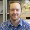 A person with short blonde hair wearing a blue flannel collared shirt is in front of metal shelves with a long-snouted skull visible on a top shelf behind them.
