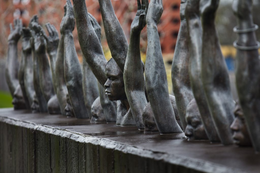 Una foto muestra una estatua de figuras de metal fundido que asemejan a varios seres humanos en fila, la mayoría de barbilla para arriba, y la del centro de busto para arriba. Todos tienen los ojos cerrados y los brazos levantados.
