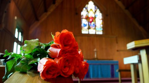 A bouquet of red roses rests on top of a wooden coffin in a church under a stained-glass window.