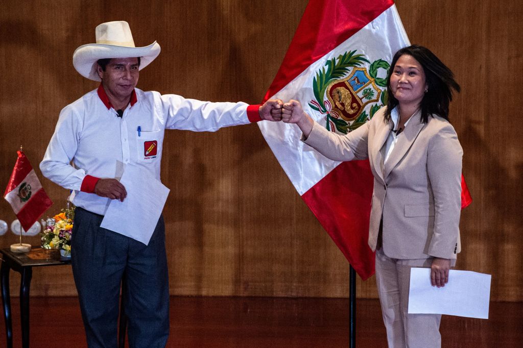 A person in a cowboy hat bumps fists with a person in a khaki pants suit in front of a red and white flag.
