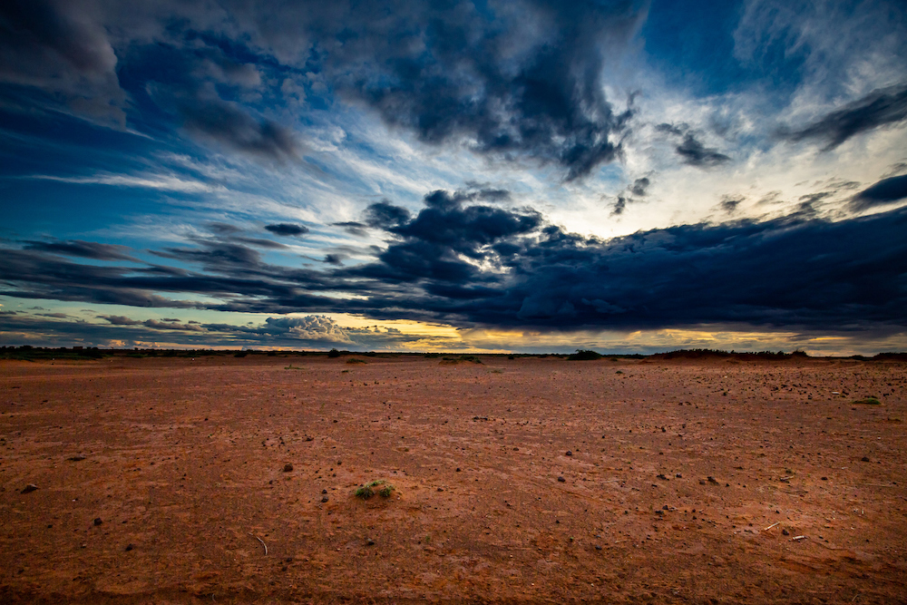 An empty sandy desert sits below a darkening sky with yellow from the sun peeking through.