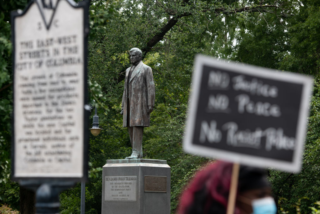In the center of the photo, a statue of a man in a long coat stands on a block embossed with a placard reading “Benjamin Ryan Tillman.” In the foreground, a protest sign reads, “No Justice, No Peace, No Racist Police.”