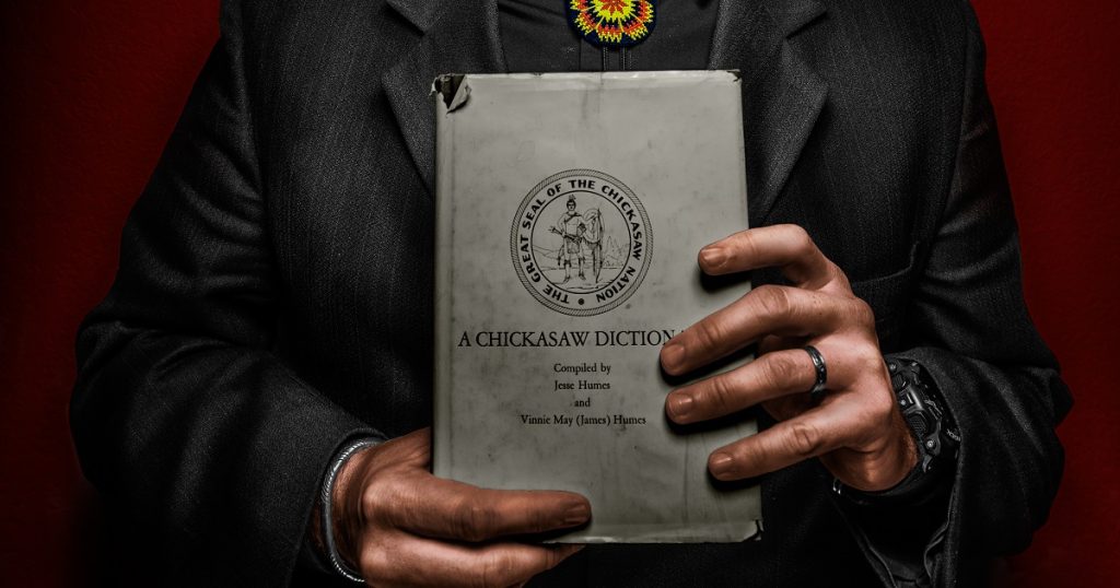 Two hands hold a book whose cover reads, "The Great Seal of the Chickasaw Nation; A Chickasaw Dictionary compiled by Jessie Humes and Vinnie May (James) Humes"