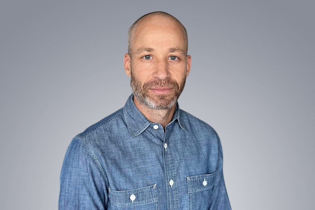 A man with a lightly shaven head and gray beard smiles into the camera.