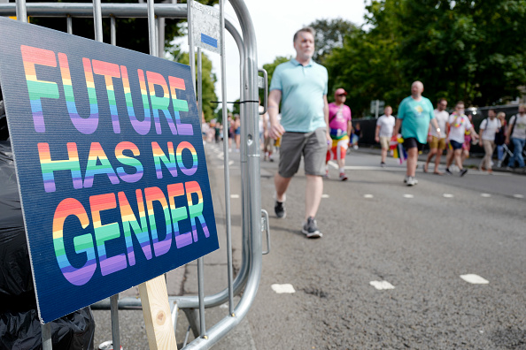 A group of people walk down a road with a large sign on their right that reads, "future has no gender" in rainbow-colored, capitalized letters.
