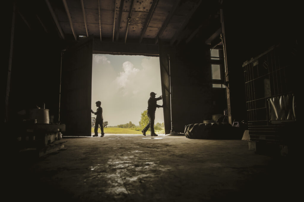 A photograph features the dark interior of a large barn and light spilling in through a doorway as an older man opens the right door and a young boy opens the left door.