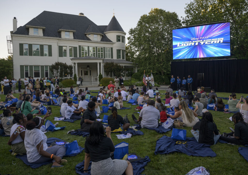 Many people sit outside on blankets on a grass field and look up at a large screen that reads Lightyear against swirling colors.