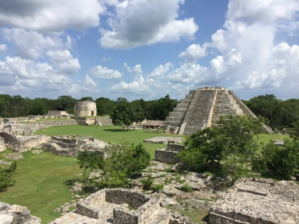 A tall, stair-step pyramid of beige stone in the distance towers over other stone ruins, with its top level plateauing just under a light-blue sky with white clouds.