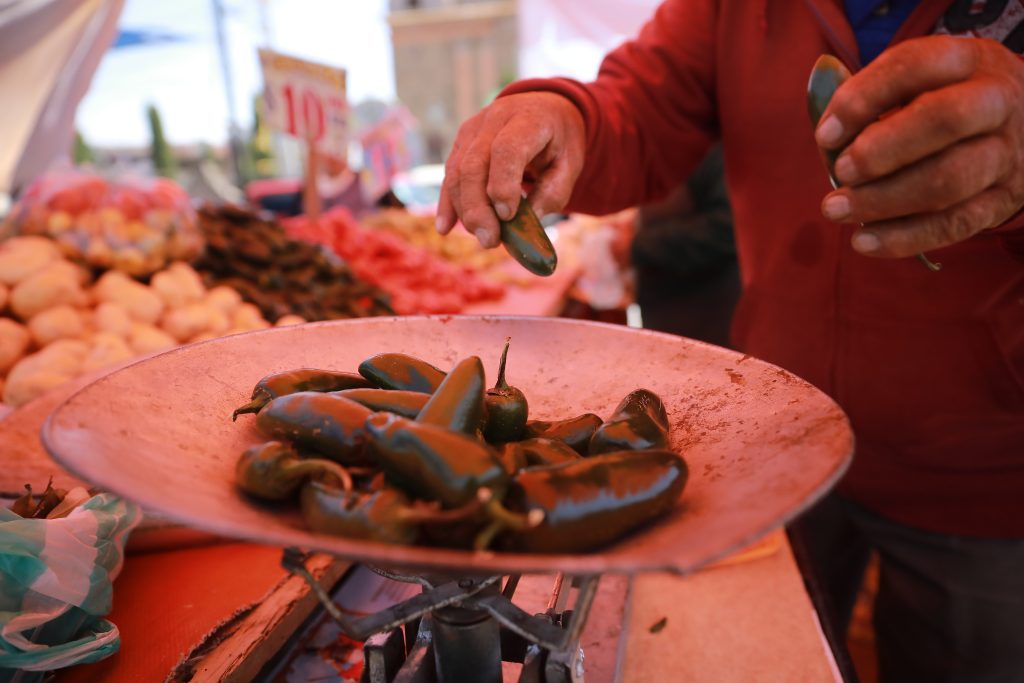 The photograph features a person’s hands placing green jalapeño peppers on a gray scale at a farmer’s market. Price tags and other produce vendors are visible in the background.