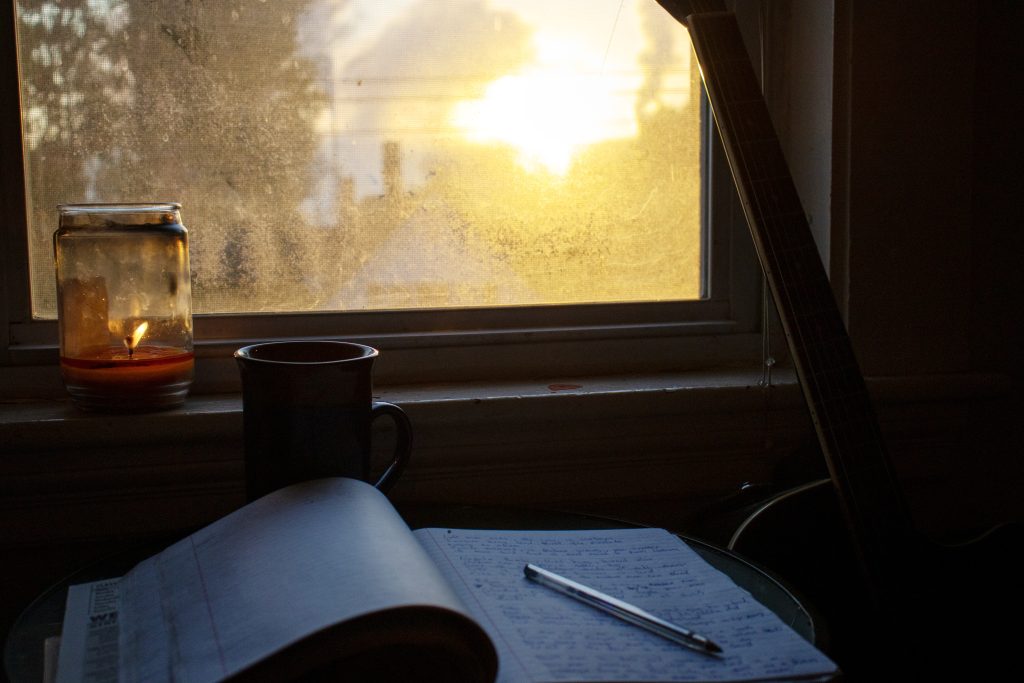 In front of a window in a dark room, a pen rests on an open notebook on a desk. Yellow sunlight shines through the window and causes a glare.