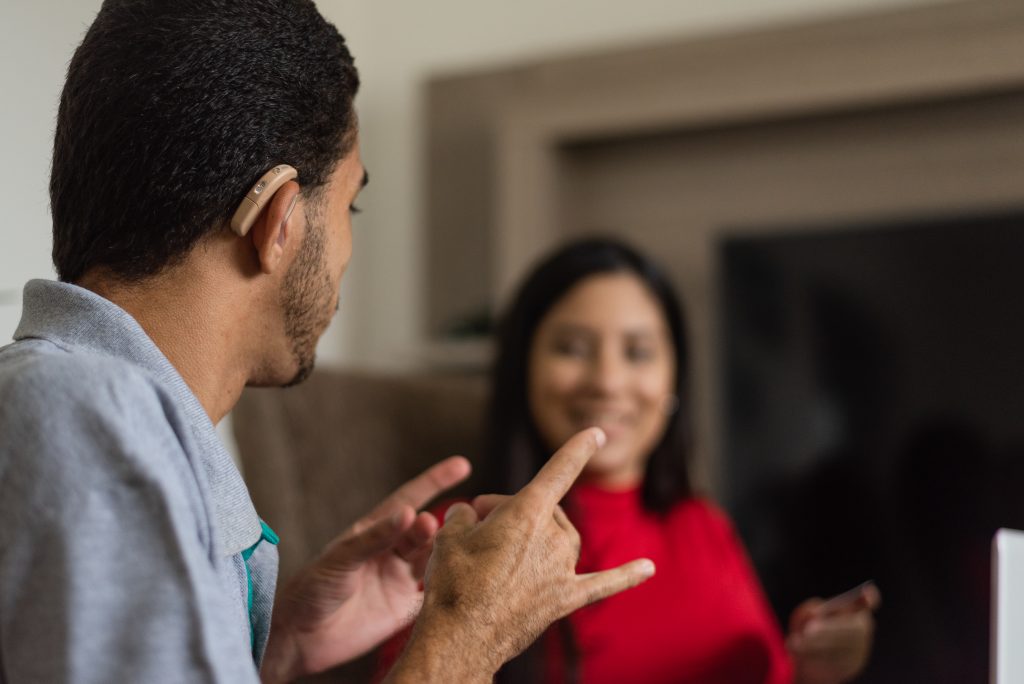 The photograph features a person with a hearing aid sitting next to a person smiling on a couch. They face each other and communicate using sign language.