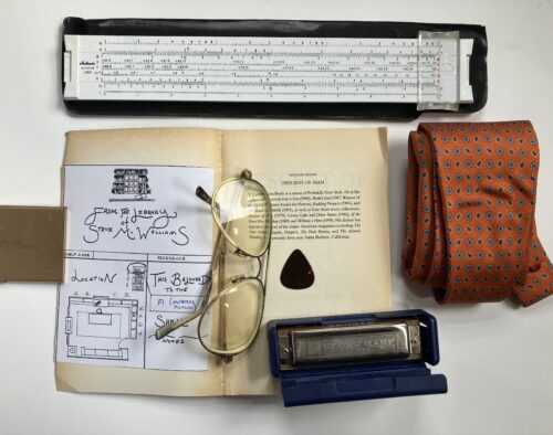 A close-up image features an open book with visible text that reads, “Descent of Man.” Around it are a pair of yellow glasses, a hand-drawn schematic, a rolled tie, a harmonica, and a slide rule.