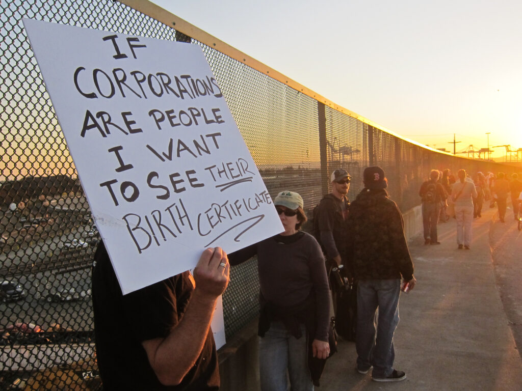 A person stands by a fence with several other people holding a sign that reads: “If corporations are people I want to see their birth certificate.”