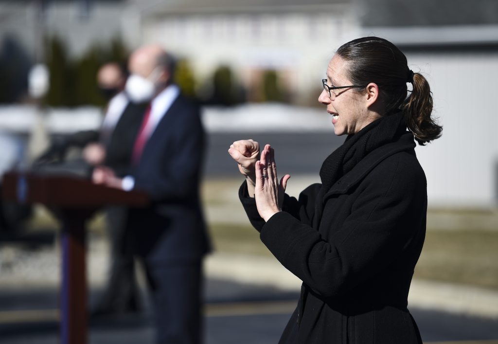 Un intérprete con gafas negras y gabardina negra gesticula en primer plano mientras un hombre con corbata roja, traje negro y mascarilla blanca está de pie en un podio al fondo.