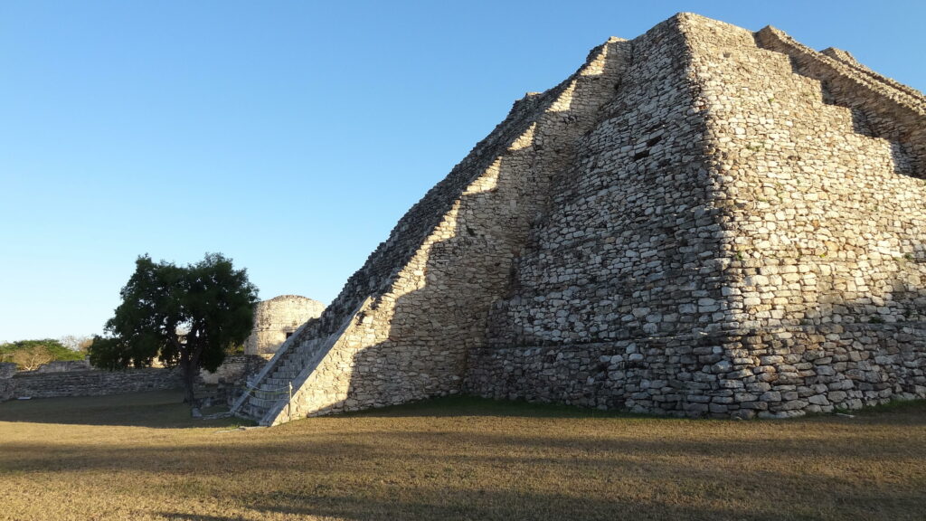 Um close-up mostra o lado sombrio de uma grande pirâmide de oito camadas feita de pedras bege com um céu azul acima e grama amarelada em sua base.