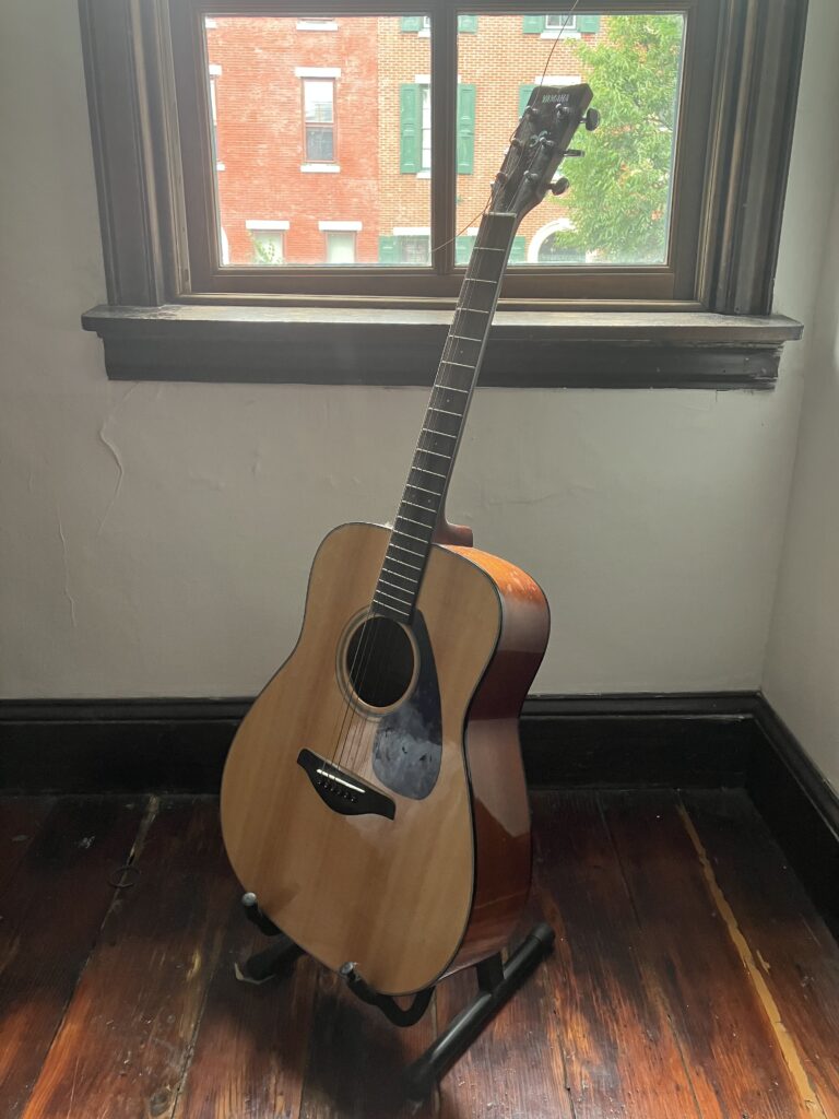 A wooden acoustic guitar rests vertically on a black guitar holder in front of a window in the corner of a room.