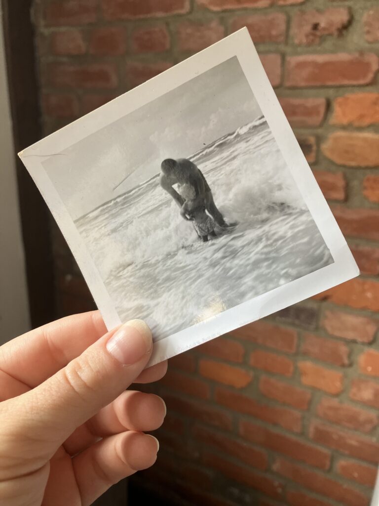 Against a brick background, a hand holds up a black-and-white photo of an adult and a child in ocean water.