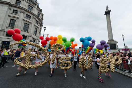 A photograph features a parade with people in the front holding up gold balloons that spell out “Disney” and those behind holding mouse-head–shaped balloons in rainbow colors.