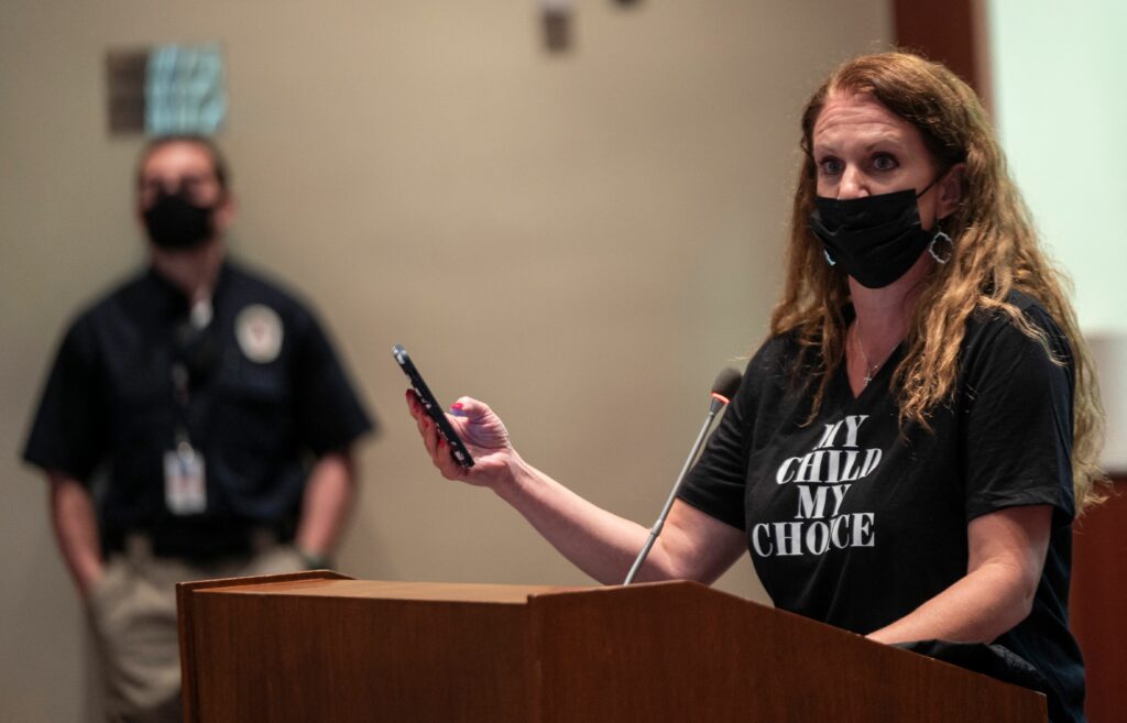 A person with long curly hair and a black face mask holds a phone and stands behind a podium. She is wearing a black T-shirt with white lettering that reads, “My child my choice.”
