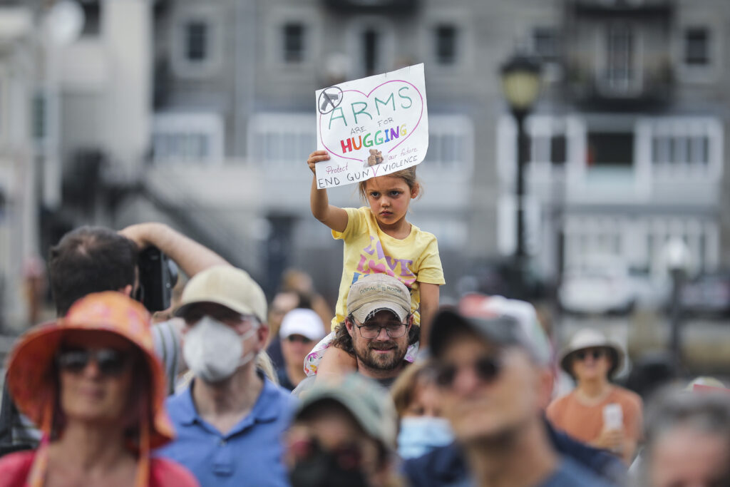 A group of people face in the same direction. A frowning child sits on a person’s shoulders and holds a multicolor handwritten sign that reads, “Arms are for hugging. Protect our Future. End Gun Violence.”
