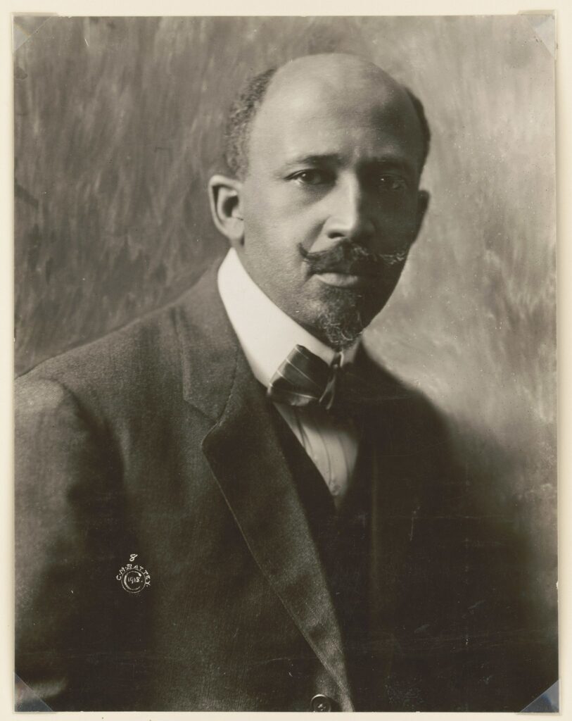A grayscale portrait features a nearly bald man with a handlebar mustache and goatee, wearing a suit jacket, white collared shirt, and pinstripe bowtie.