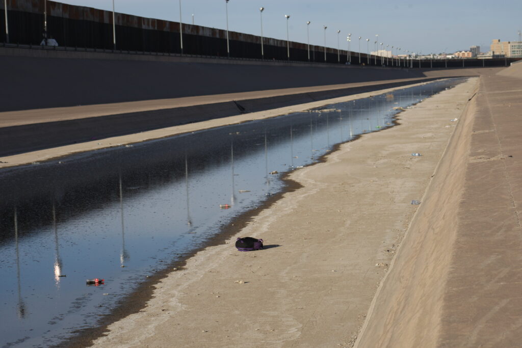 A long body of water separates a tall wall lined with streetlamps from a black backpack that rests alone on a concrete bank.