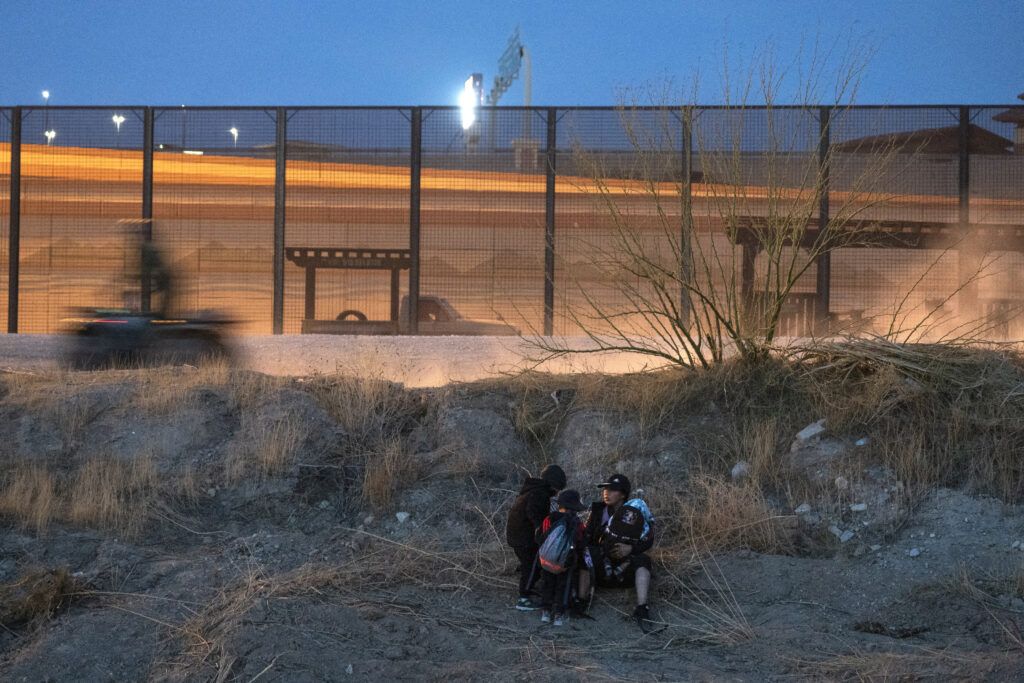 An adult and three children huddle by a grassy hill in the dark with a tall wire fence in the background.