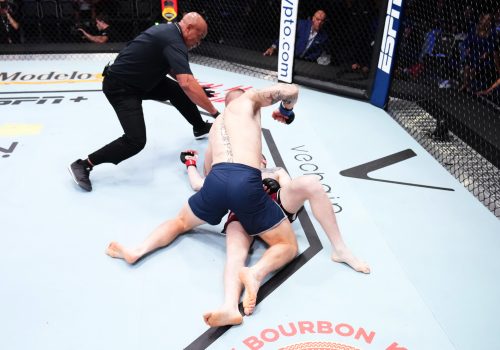 One MMA fighter pounds another fighter who is limp, laying flat on the mat, while a referee attempts to intervene.