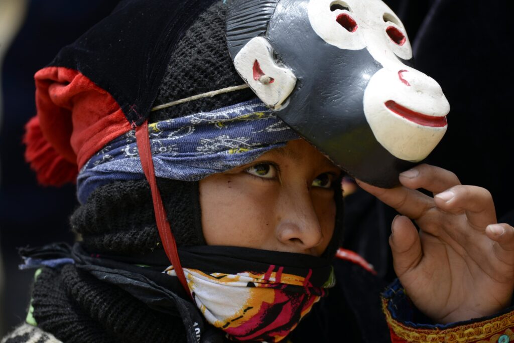 A close-up image features a child wearing a black balaclava and multicolor cloth mask. They are lifting up a character mask that resembles a monkey’s face to reveal their own.