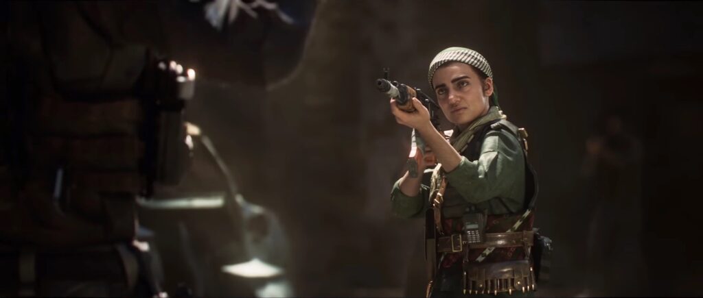 A still from an animated video game features a woman in a green uniform, headscarf, and bullet belt pointing a gun at a faceless person dressed in black in the foreground.