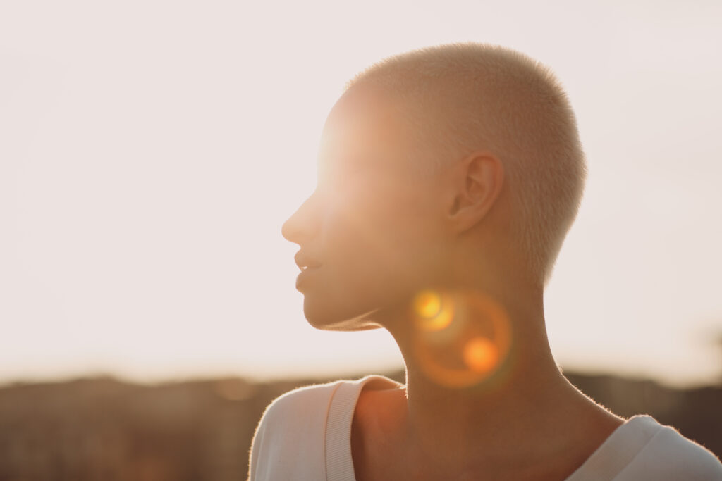 A short-haired person turns their head to their right in front of the horizon, a sun glare shining from behind them.