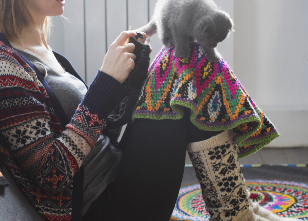 A person wearing a crocheted cardigan crochets while balancing a gray kitten on a multicolor crochet piece on their knees.