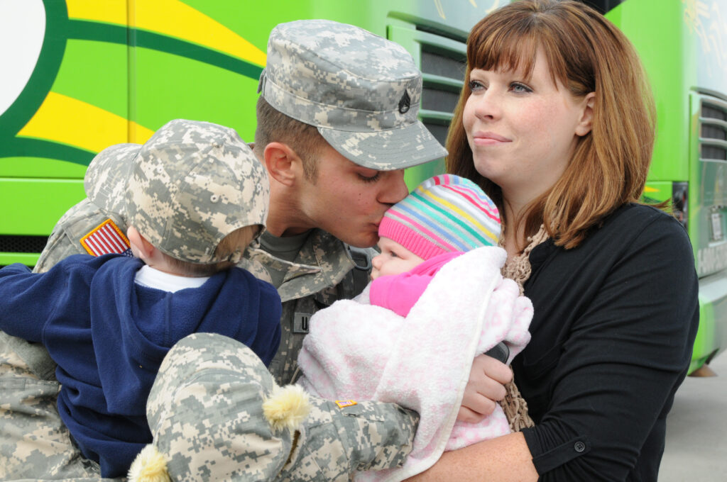 A man in military camo fatigues hugs three small children: two in camo print outfits and one in pink. A woman in a black shirt hugs them all from his left side while staring off over their heads.