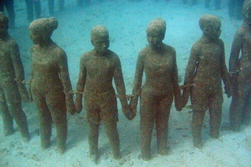 An underwater scene features statues of people standing in a circle with adjacent figures shackled together by the wrists. They are facing outward and holding hands.