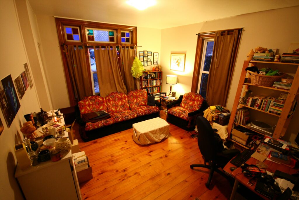This photograph features a living room with wooden floors, a sofa, an armchair, a bookshelf, a computer chair, windows, and a desk lamp casting a yellowish hue.