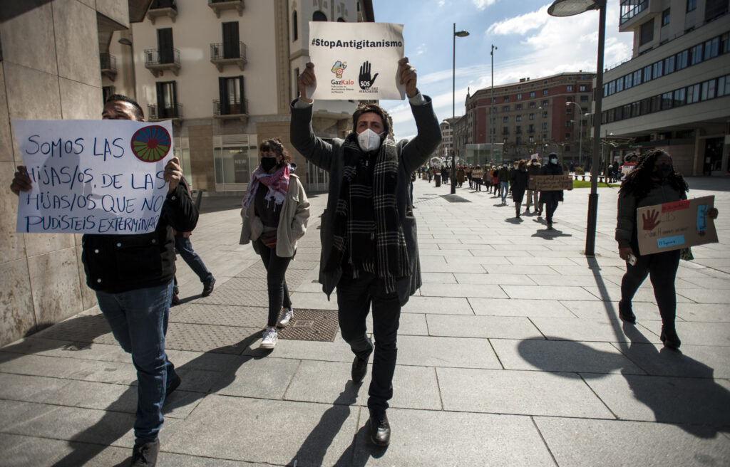 Protestors march along a city street carrying signs, one of which reads #StopAntigitanismo.