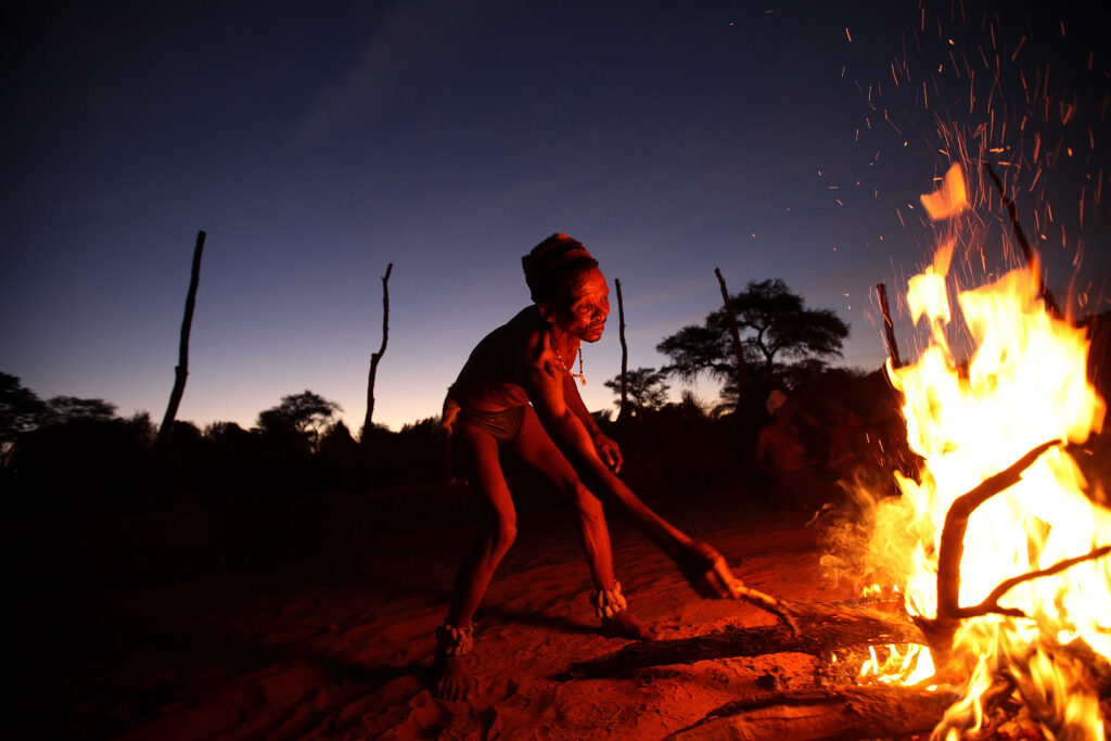 Outside in relative darkness, a person in a headwrap, loincloth, and neck and ankle jewelry stokes a giant fire with a branch.