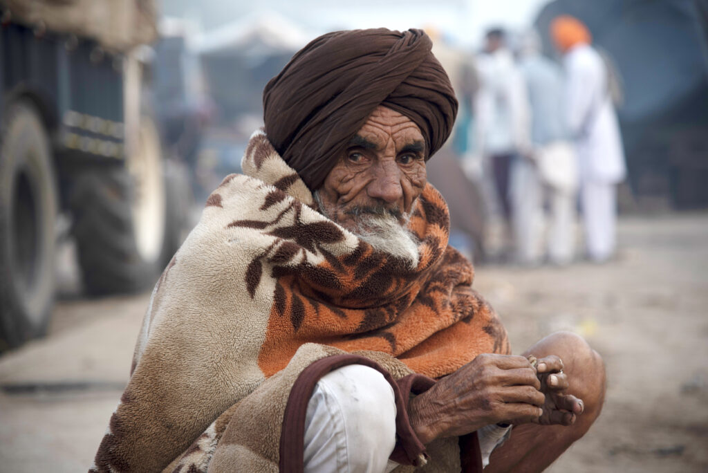 An older person with wrinkled skin, a head turban, and scarf crouches and looks into the camera. A cluster of people and a large produce truck are in the background.