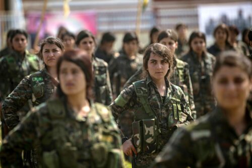 A photograph features a group of women dressed in green camo uniforms looking toward their right with stern expressions on their faces.