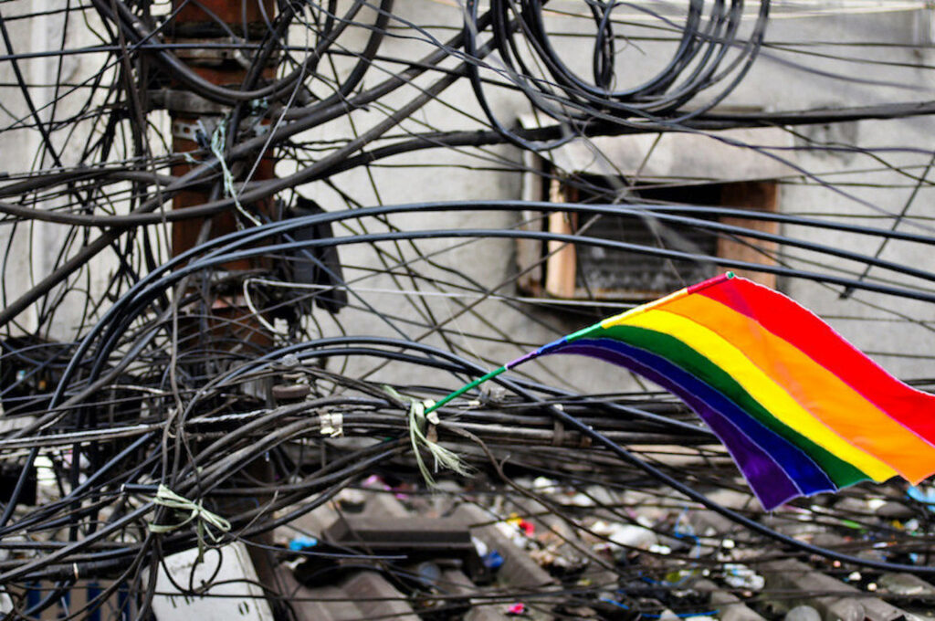A photograph features a flag with rainbow-colored stripes lodged in a tangle of wires between a telephone pole and gray tin roof with trash strewn about.