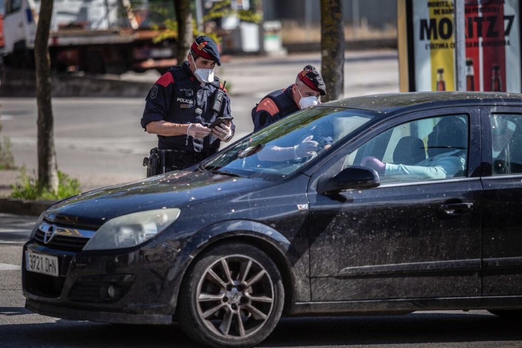Two police officers wearing face masks lean over a black car to speak with the driver.