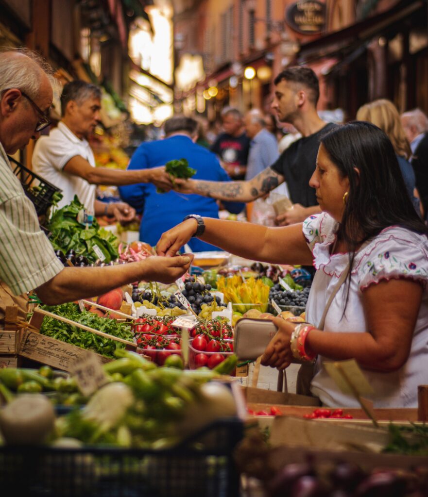 People pay for fruits and vegetables in a farmers market filled with red, yellow, and green produce.