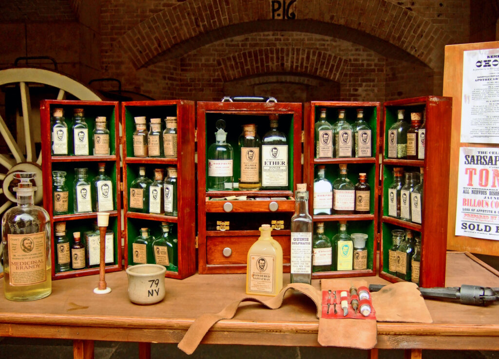 A wooden medicine cabinet on a wooden table displays numerous clear and green medicine bottles.