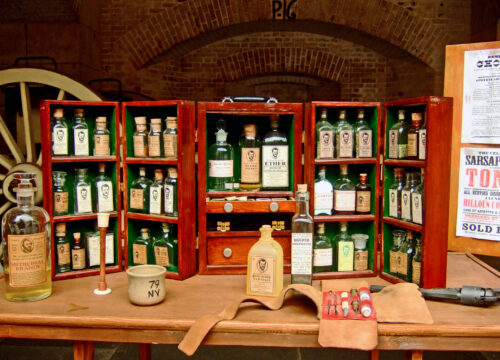 A wooden medicine cabinet on a wooden table displays numerous clear and green medicine bottles.