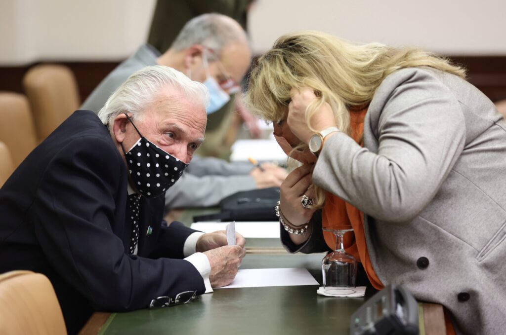 A man with gray hair and a polka-dot face mask sits at a table and listens to a person leaning over and speaking with him.