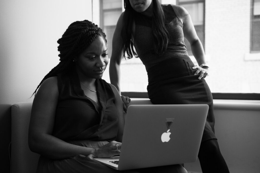 One woman with braids sits at a laptop, another woman standing over her shoulders. Both look at the screen.