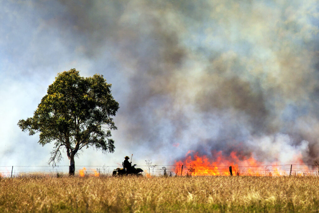 A photograph features a yellow grassy field in the foreground and large orange flames, smoke-covered blue sky, person on a tractor, and lone green leafy tree in the distance.